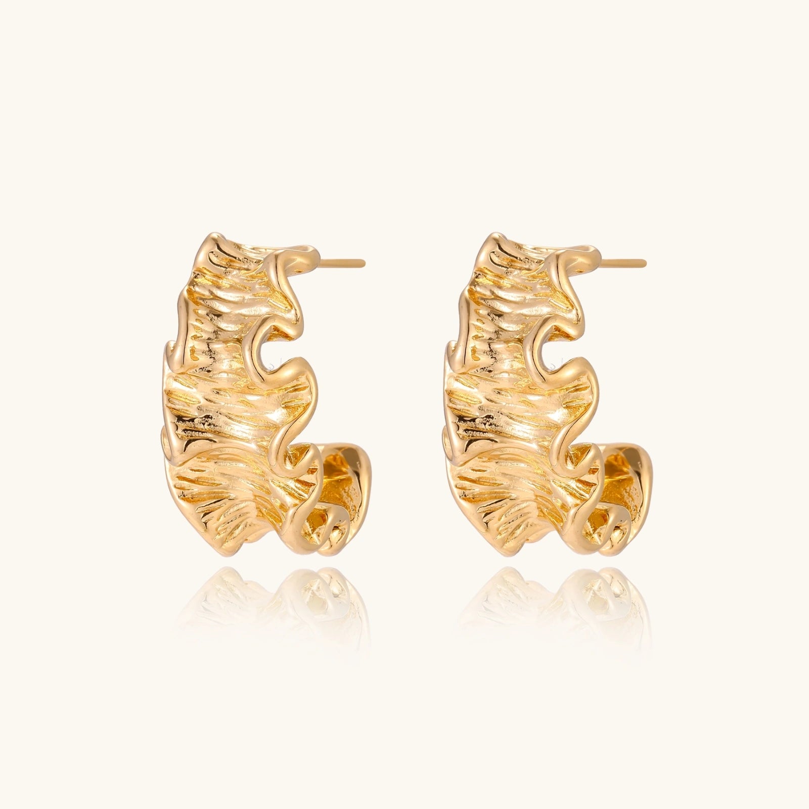 Pair of gold huggie hoops with sculpted wave texture and dimensional ripples on neutral background.